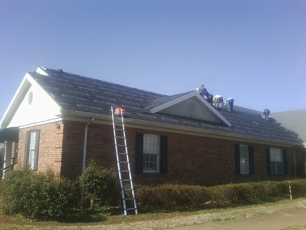 Crew tearing shingles off a brick ranch roof with a ladder set — Metal Roof Repair work by our crew in Crane Hill
