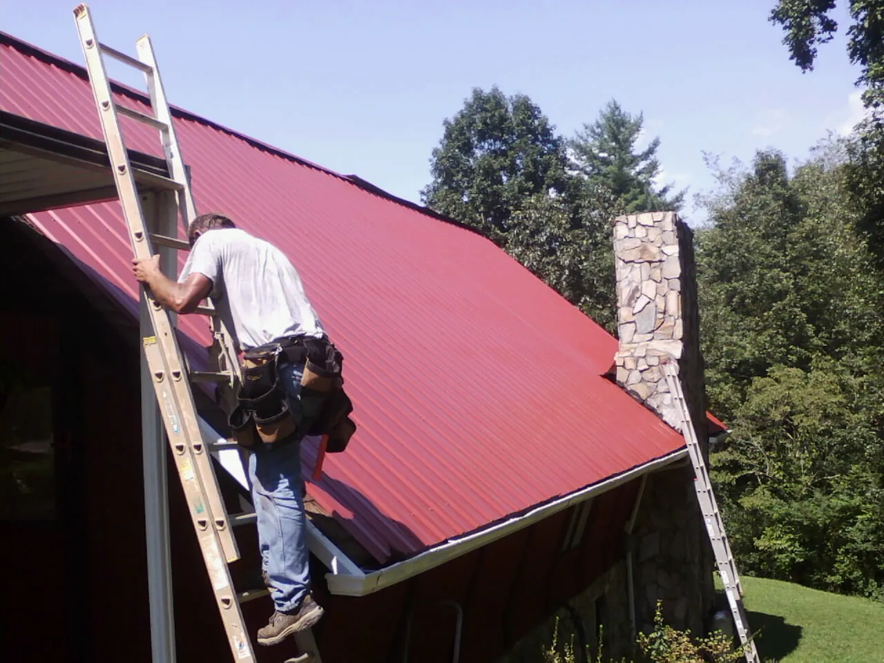 Roofer on a ladder beside a red metal roof and stone chimney — Soffit & Fascia Repair by Roofing Brothers in Crane Hill